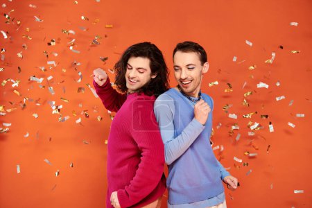 Foto de Feliz bien parecido gay amigos en elegante ropa con maquillaje posando bajo confeti lluvia, orgullo - Imagen libre de derechos