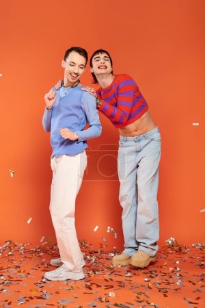 Téléchargez les photos : Deux gays élégants joyeux en tenue vibrante s'amusant sous la pluie de confettis sur fond orange - en image libre de droit