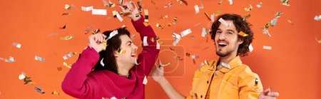 Téléchargez les photos : Joyeux élégant gay les hommes dans vibrant attires avoir amusant sous confettis pluie sur orange toile de fond, bannière - en image libre de droit