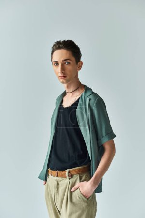 A young man proudly poses in a green shirt and tan pants, showcasing his vibrant queer fashion in a studio setting.