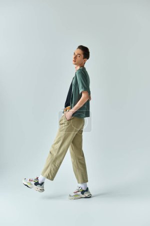 Photo for A young queer person confidently poses in a studio, wearing a stylish t-shirt and khaki pants on a grey background. - Royalty Free Image