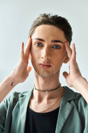 Ein junger queerer Mensch steht mit den Händen auf dem Kopf, tief in sich versunken, in einem Studio mit grauem Hintergrund.