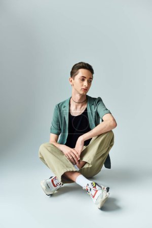 A young queer person sits on the floor, wearing a green jacket and sneakers, exuding confidence and pride in a studio setting.