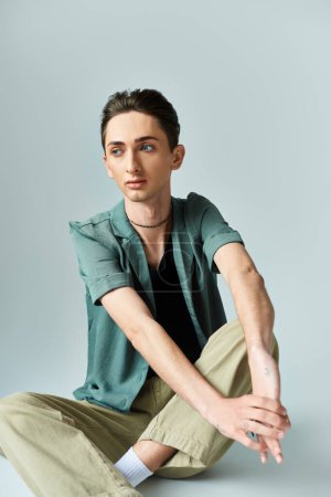 A young queer person, wearing a green shirt and tan pants, sits on the floor in a reflective pose against a grey background.