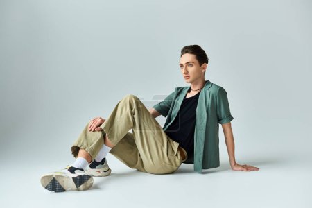 Ein junger queerer Mensch in braunem Hemd und khakifarbener Hose sitzt in nachdenklicher Pose auf dem Boden in einem Studio vor grauem Hintergrund..