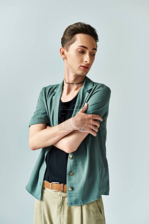 A young queer person confidently poses in a studio wearing a green shirt and tan pants, expressing LGBT pride on a grey background.