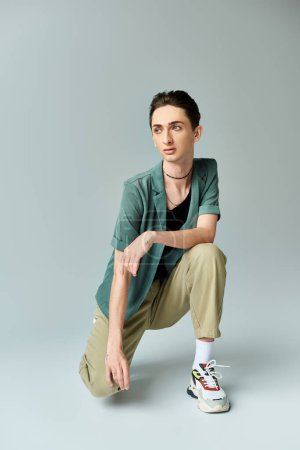 A young queer person crouching down in a pose of contemplation against a grey background in a studio setting.