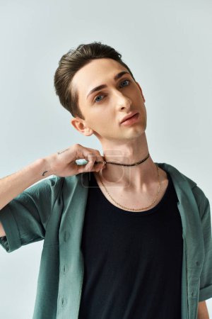 Una persona queer joven toma una postura segura en un ambiente de estudio, vistiendo una camisa negra y un collar de gargantilla.