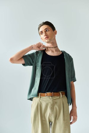 A young queer person wearing a green shirt and tan pants poses in a studio on a grey background, embodying lgbt and pride.