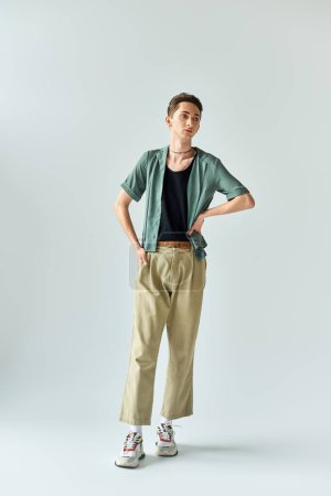 Young queer person confidently posing in studio wearing a tan shirt and khaki pants on a grey background.