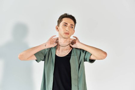 A young queer person confidently poses in a studio wearing a green shirt against a grey background.
