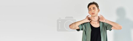 Eine junge queere Person posiert selbstbewusst mit den Händen auf dem Kopf in einem Studio vor grauem Hintergrund.
