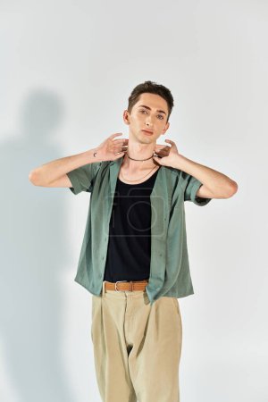 Une jeune queer dans un studio, vêtue d'une chemise verte et d'un pantalon bronzé, exsudant fierté et confiance dans un contexte gris.