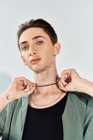 A young queer man adjusts his necklace, showcasing pride and elegance in a studio setting against a grey background.
