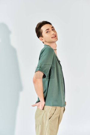 A young queer person confidently poses in a studio wearing a green shirt and khaki pants, standing out against a grey background.