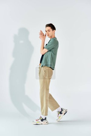 A young queer person in a green shirt and khaki pants stands confidently in front of a white wall in a studio setting.