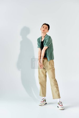 A young queer person strikes a stylish pose in a tan shirt and khaki pants, exuding confidence in front of a white background.