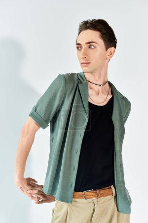 Ein junger queerer Mensch posiert selbstbewusst in einem Atelier mit grünem Hemd und brauner Hose vor grauem Hintergrund.
