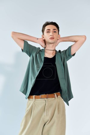 Eine junge queere Person posiert selbstbewusst in einem Studio-Setting, trägt ein stylisches grünes Hemd und eine khakifarbene Hose vor grauem Hintergrund.