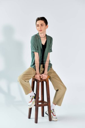Ein junger queerer Mensch sitzt auf einem Hocker in einem Atelier und posiert vor grauem Hintergrund mit einem Gefühl von Stolz und Authentizität.