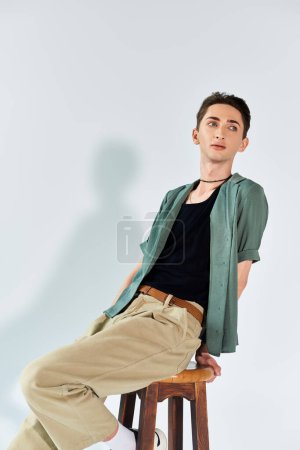 A young queer person in a green shirt and khaki pants sits thoughtfully on a stool in a studio against a grey background.