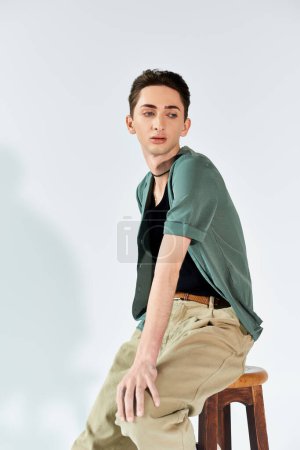 A young man confidently sits on a stool in a stylish green shirt and tan pants against a grey studio backdrop.