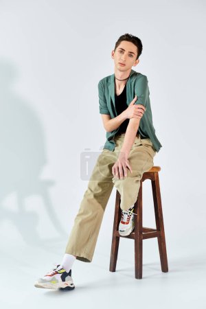 A young queer person confidently sits on a stool in a studio, exuding pride and empowerment against a grey background.