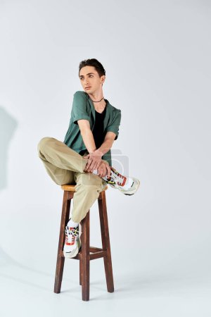 Eine junge queere Person sitzt elegant auf einem Hocker in einem Atelier und zeigt Zuversicht und Stolz.