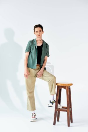 Téléchargez les photos : Un jeune queer élégant se tient avec confiance sur un tabouret portant une chemise verte et un pantalon kaki dans un studio sur fond gris. - en image libre de droit