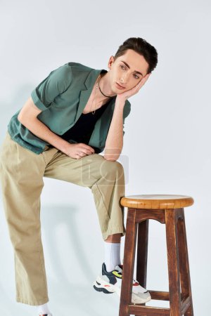 queer person, part of the LGBTQ+ community, sits proudly on a stool in khaki pants and a green shirt against a grey background.