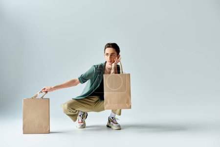 A young queer crouches, exuding pride, holding shopping bags in a studio against a grey background.