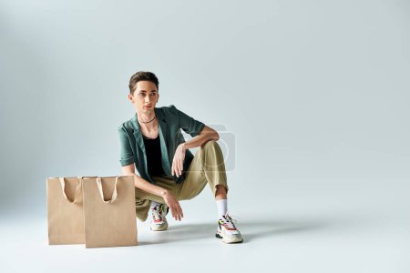 Photo for A young queer person sitting on the floor surrounded by colorful shopping bags in a studio on a grey background. - Royalty Free Image