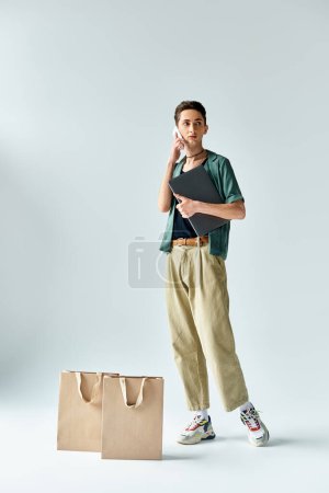 Ein stylischer junger Mann balanciert mühelos zwischen Einkaufstaschen und Handy und strahlt vor grauem Hintergrund Selbstbewusstsein und urbanes Flair aus..