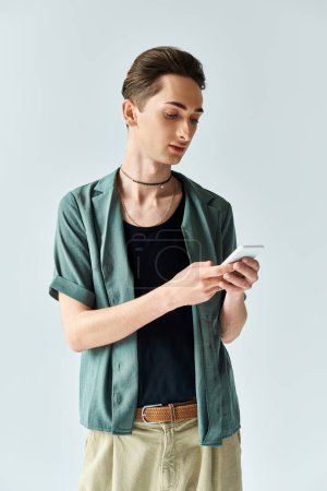 A stylish young queer man, wearing a green shirt and tan pants, checks his phone against a grey studio backdrop.