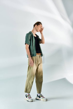 A young queer person in a tan shirt and khaki pants stands confidently against a plain white backdrop.