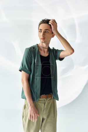 Ein junger queerer Mensch mit Stolz trägt ein grünes Hemd und eine khakifarbene Hose und posiert selbstbewusst in einem Studio vor grauem Hintergrund.