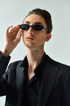 A young queer person strikes a confident pose in a sharp suit, wearing sunglasses on a grey background.