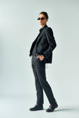 A young queer person exudes confidence in a black suit and sunglasses, striking a pose in a stylish studio setting.