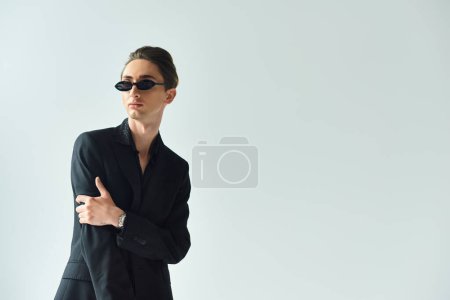 Young queer person with black suit confidently stands with arms crossed in studio set against grey background.