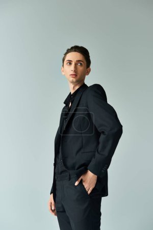 Un joven queer toma una postura confiada en un elegante traje negro contra un fondo gris, exudando orgullo y estilo.