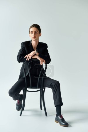 A young queer person confidently sits in a suit on a chair against a grey background, exuding pride and empowerment.