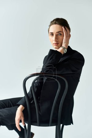 A young queer person exuding confidence, sitting on a chair in a stylish black suit against a grey studio background.