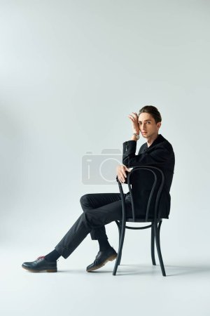 Ein stilvoller junger Mann im Anzug sitzt auf einem Stuhl und strahlt Zuversicht und Besinnlichkeit in einem Studio mit grauem Hintergrund aus..