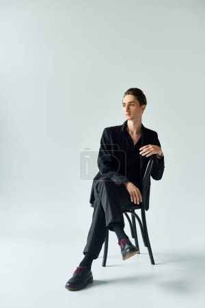 Foto de Un joven con un traje negro emana confianza mientras está sentado en una silla en un estudio sobre un fondo gris. - Imagen libre de derechos