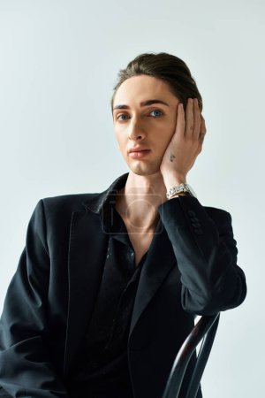 A young queer person in a stylish black suit sits on a chair against a grey backdrop.