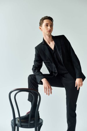 Foto de A stylish young man, dressed in a sleek black suit, strikes a pose while sitting on a chair in a studio with a grey background. - Imagen libre de derechos
