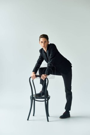 Un jeune queer en costume stylé se penche en toute confiance sur une chaise dans un décor de studio avec un fond gris.