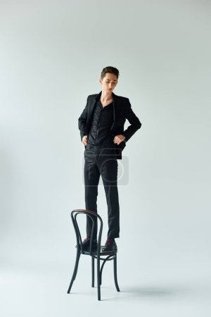 Stylish queer man in a black suit standing confidently on a chair in a studio setting, showcasing pride and elegance.