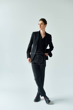 Un joven queer, vestido con un elegante traje negro, posa con confianza para la cámara en un estudio con un fondo gris.