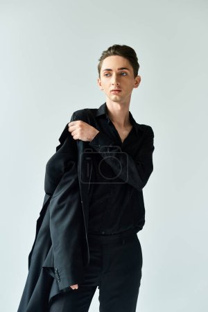 A young queer man confidently poses in a stylish black suit in a studio setting against a grey background.